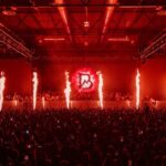 Flame effects and red stage lighting over a packed crowd at an industrial warehouse techno event