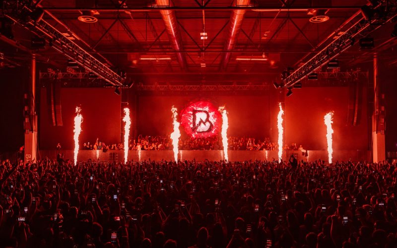 Flame effects and red stage lighting over a packed crowd at an industrial warehouse techno event